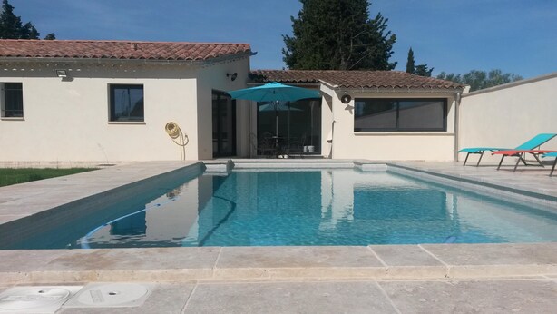 Une piscine avec une terrasse carrelée, un patio couvert avec des chaises, et un bâtiment doté d’un toit en tuiles.