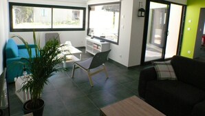 Un salon moderne avec un canapé noir, un bureau blanc et une chaise bleue. Une grande fenêtre offre une vue sur l’extérieur.