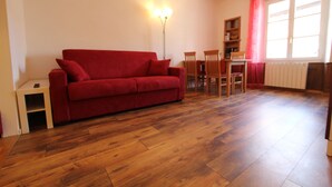A living room with a red sofa, wooden flooring, a dining table with chairs, a floor lamp, and a window with a red curtain.