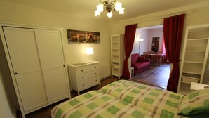 A bedroom with a bed, a nightstand, a dresser, a painting on the wall, and a view of a dining area through a door.