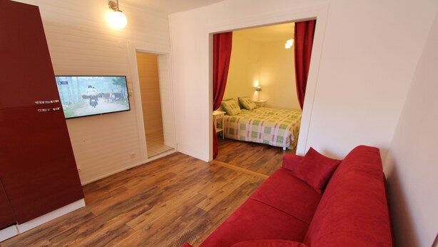 A room with a red sofa, wooden flooring, a TV on the wall, and a bed with a checkered blanket.