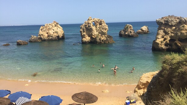 Une plage à l’eau turquoise et limpide, bordée de formations rocheuses et ponctuée de parasols.