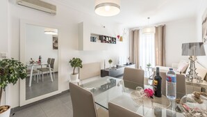 Un salon moderne avec une table à manger en verre, un canapé et une table basse. Un téléviseur est fixé au mur, ainsi qu'une bibliothèque et une plante en pot.