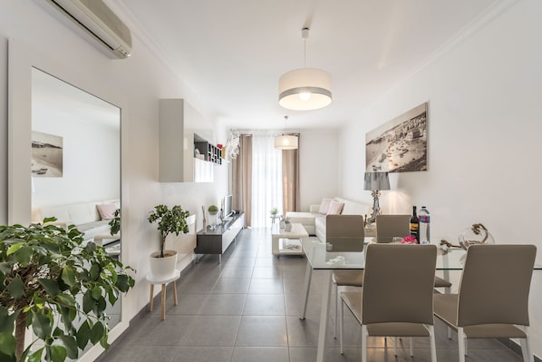A modern living room with a glass coffee table, beige sofas, and a dining area with chairs.