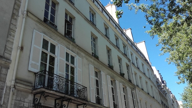 A multi-story building with white shutters, balconies with wrought iron railings, and a clear blue sky in the background.