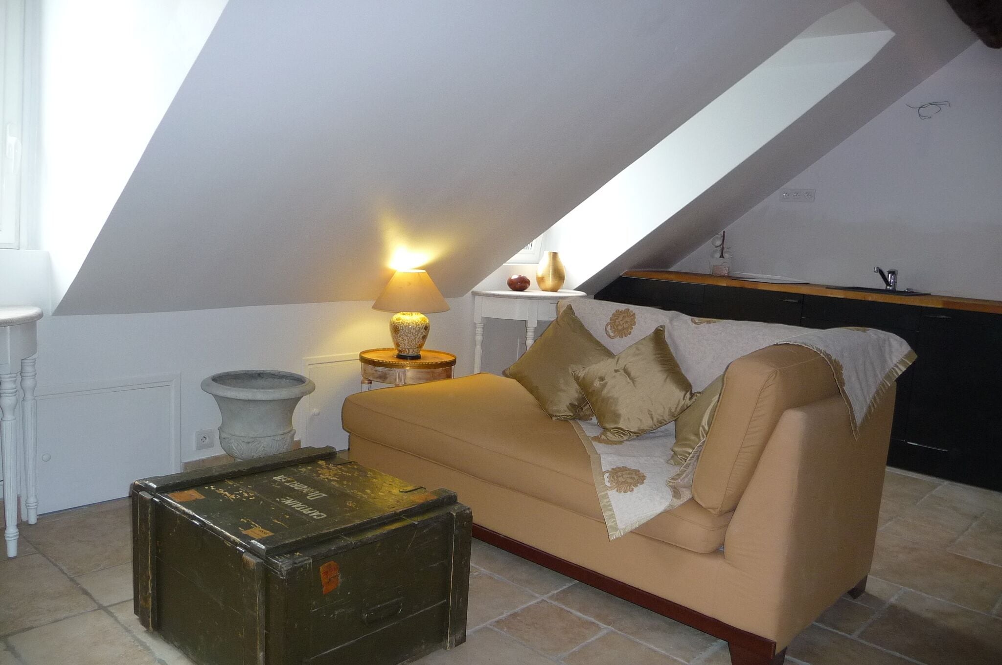 Une chambre à coucher confortable, aménagée en mezzanine avec un plafond en pente, un canapé beige, une petite table avec une lampe et une malle ancienne.