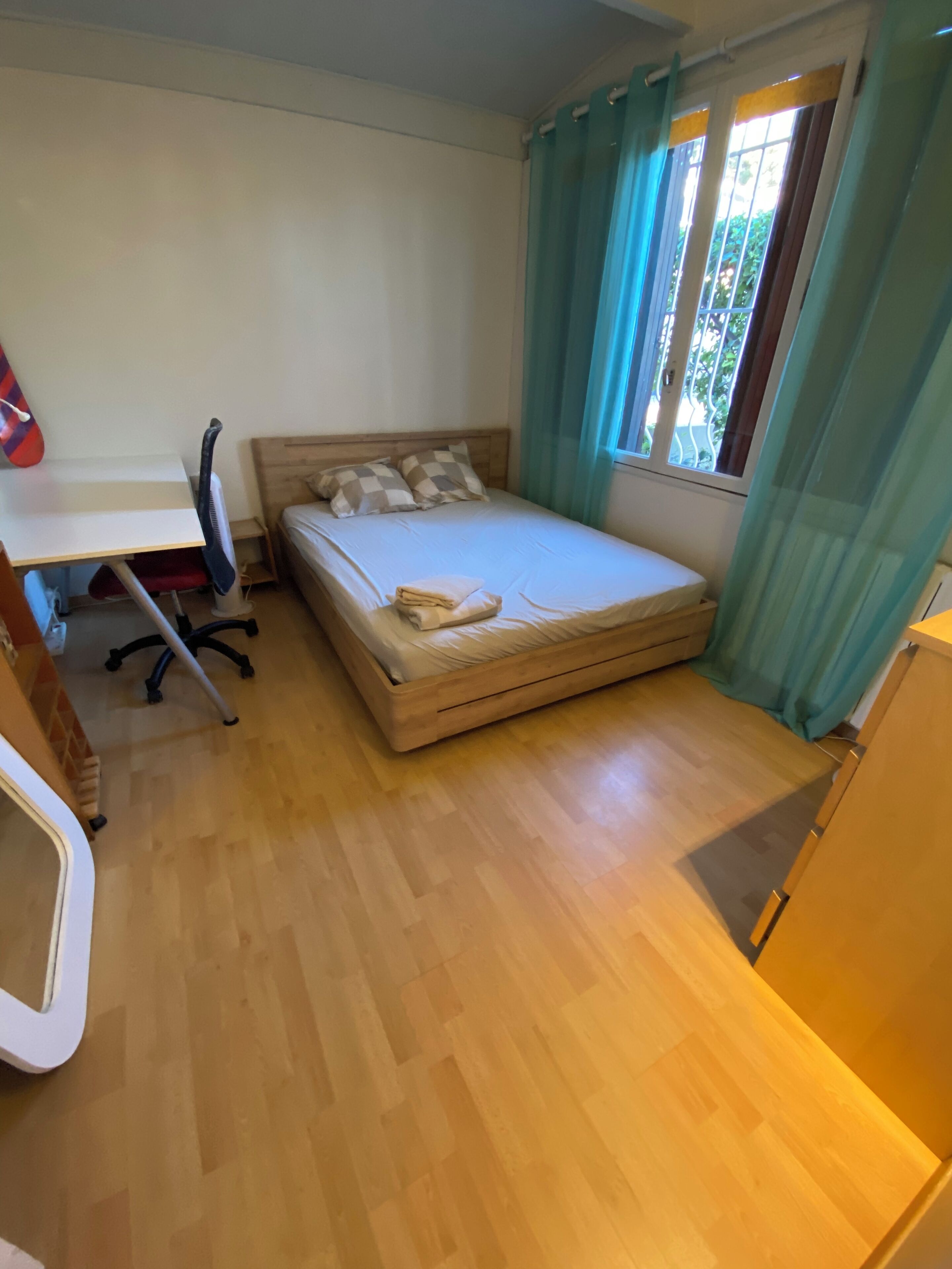 Quarto compacto com uma cama de solteiro, uma escrivaninha com cadeira e uma janela com cortinas.