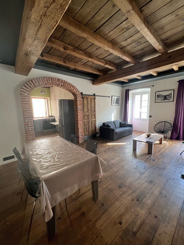 A spacious room with a wooden ceiling, a brick archway, a dining table with chairs, a sofa, and a small table with a bowl.