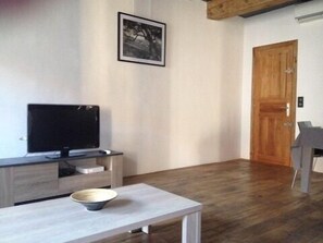 A modern living room with a flat-screen TV on a wooden stand, a dining table with a bowl, and a wooden door.