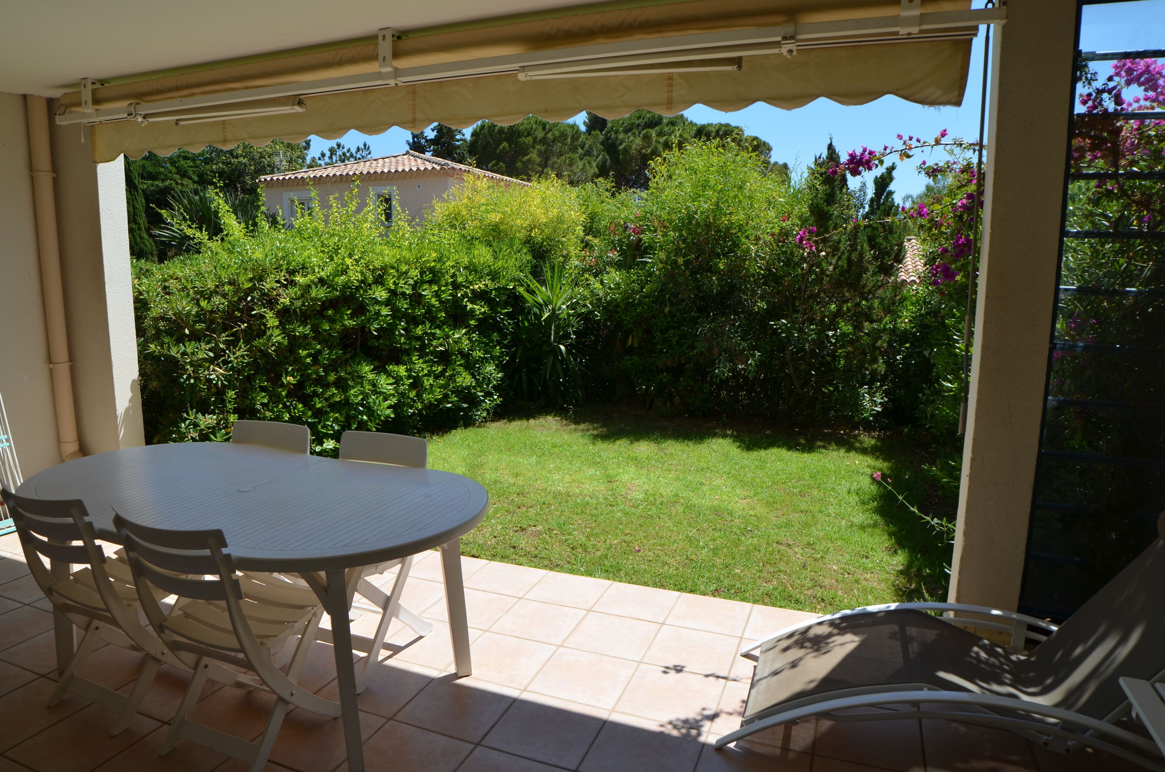 A covered patio with a table and chairs, a view of a garden with various plants and a house in the background.