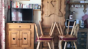 Un salon chaleureux avec un meuble en bois, deux fauteuils en bois ornés de motifs en forme de cœur, un téléviseur et une bibliothèque remplie de livres.