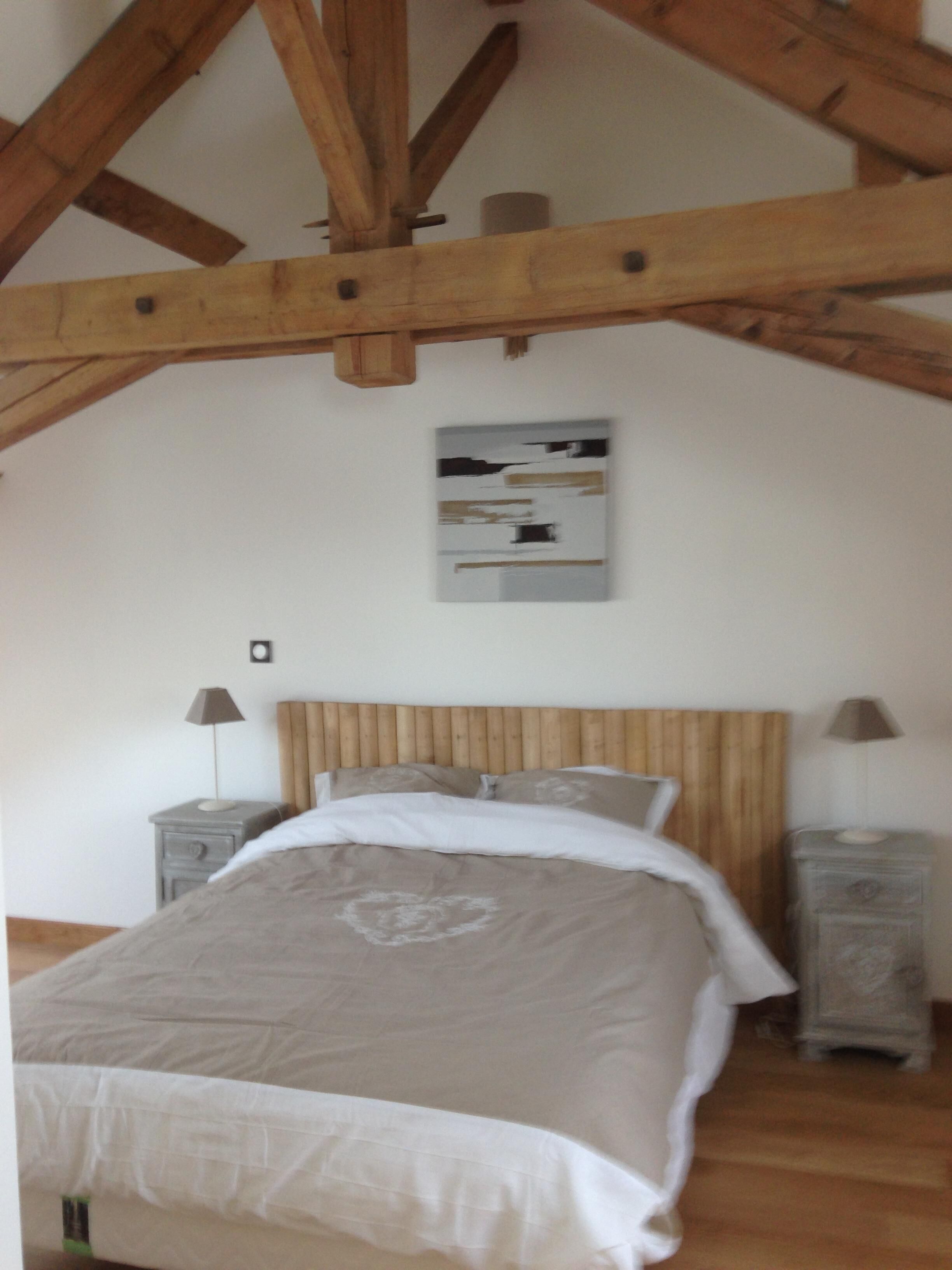 Een slaapkamer met een houten plafond, een bed met een wit-beige dekbedovertrek, twee nachtkastjes met lampen, en een schilderij aan de muur.