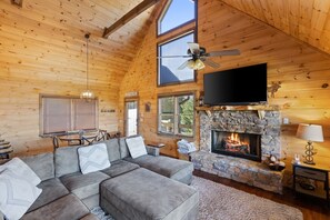 A log cabin interior with a stone fireplace, a large sofa, a ceiling fan, and a TV mounted on the wall.