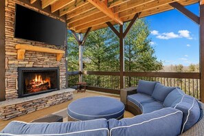 A stone fireplace, outdoor seating, and a wooden deck with a view of trees.