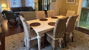 Un coin repas avec une table blanche et des chaises en osier, un lustre et une vue sur le salon.