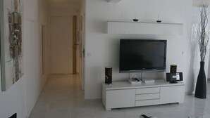 Ein modernes Wohnzimmer mit einem Flachbildfernseher auf einer weißen Unterhaltungsecke, einer schwarzen Vase mit Zweigen und einem Flur, der in ein weiteres Zimmer führt.