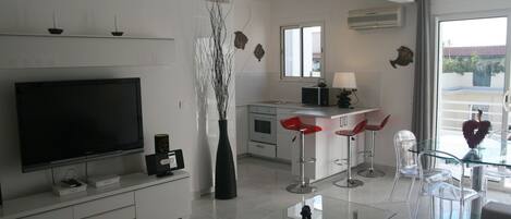 A modern living room with a flat-screen TV, a glass coffee table, and a dining area with red bar stools.