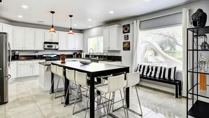 A modern kitchen with white cabinets, a central island with chairs, pendant lighting, and a window with blinds. - Buddha Estate, Pool , Spa, Sauna, Game Room (Las Vegas)