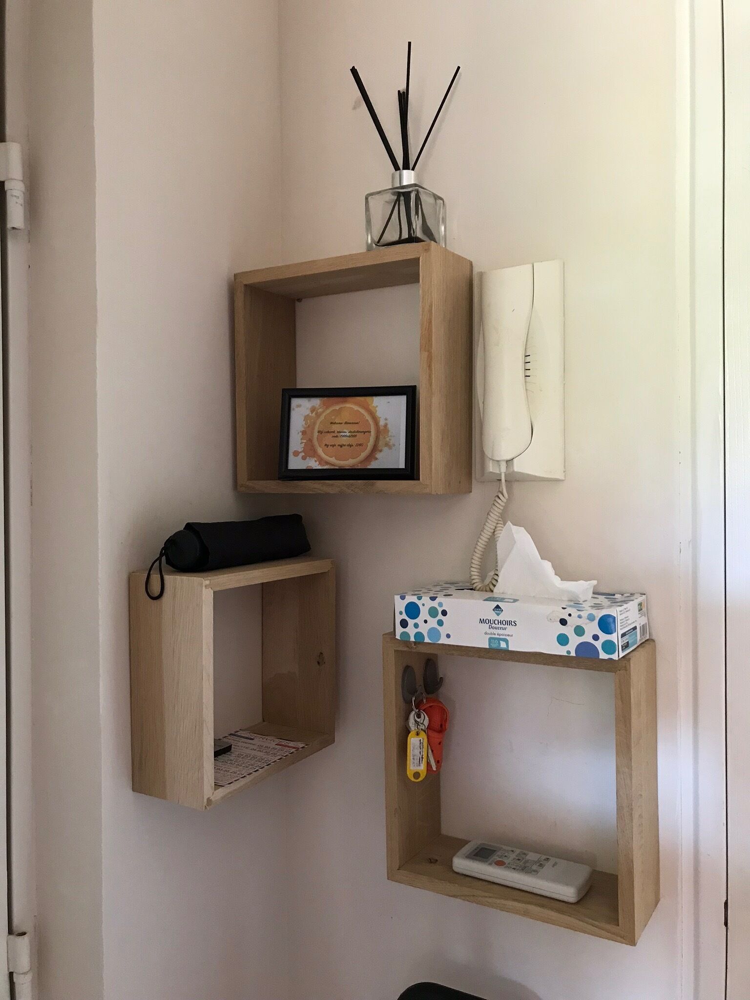 A corner wall with three wooden shelves. The top shelf holds a framed certificate and a reed diffuser. The middle shelf has a white telephone and a box of tissues. The bottom shelf contains a remote control and a keychain.