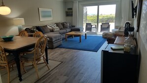 A living room with a sofa, coffee table, dining table, chairs, and a view through a glass door to the outdoors.