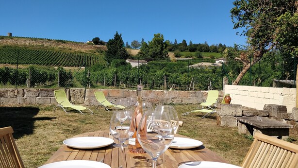 Une table en bois, installée à l’extérieur, est dressée avec des verres à vin et des assiettes, entourée de chaises vertes. On aperçoit en arrière-plan un vignoble et un mur de pierres.