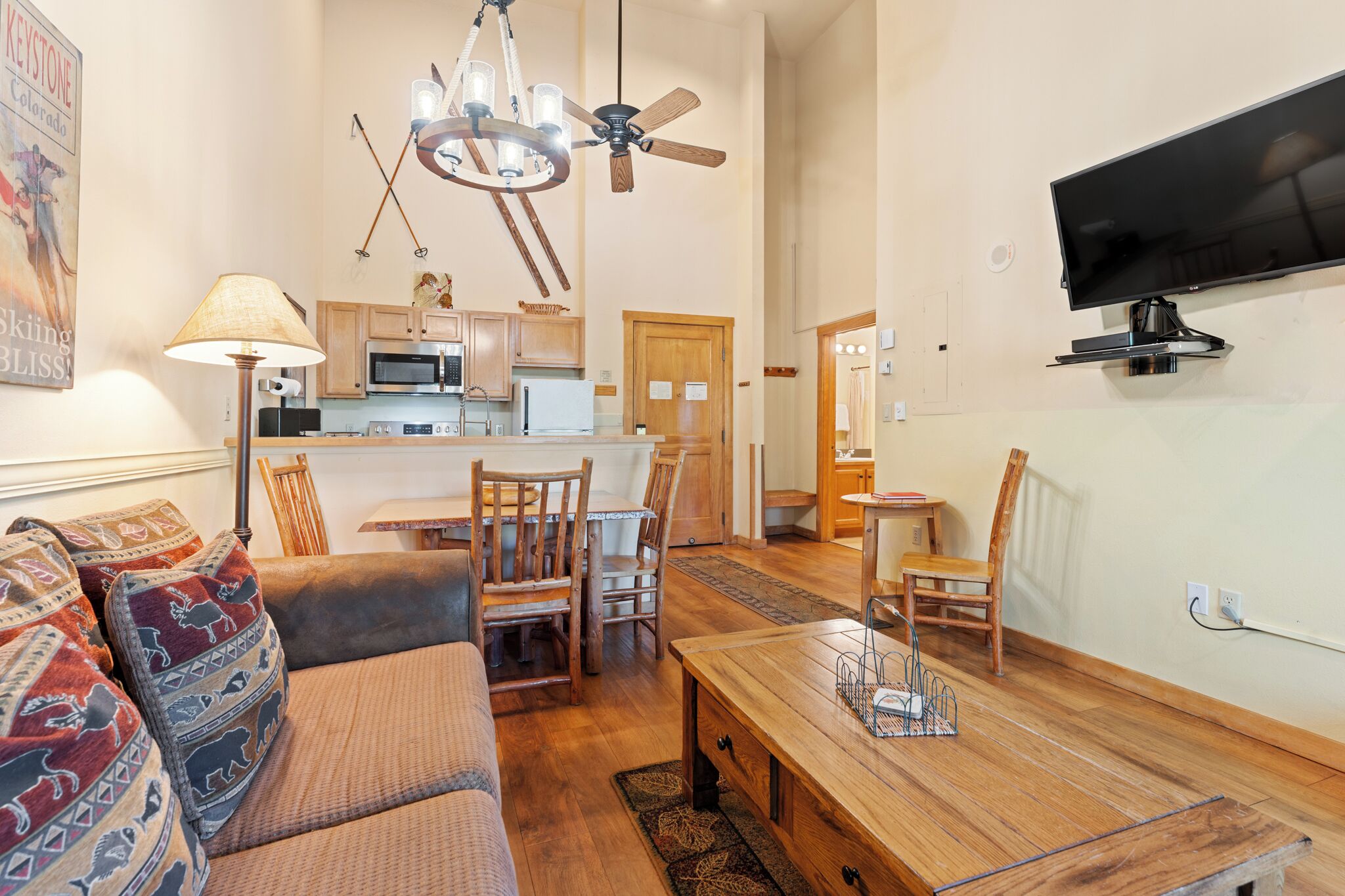 Un salon comprenant un canapé, une table à manger, des chaises, un lustre, un ventilateur de plafond, un téléviseur et un coin cuisine.
