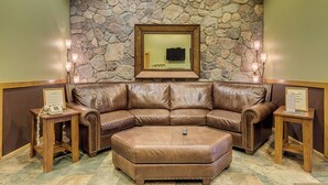 Un canapé et un pouf en cuir marron se trouvent dans un hall d’entrée, avec un mur en pierre en arrière-plan et un téléviseur encadré.