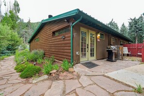 Uma cabana de toras com telhado verde, uma porta da frente e uma churrasqueira na área externa.