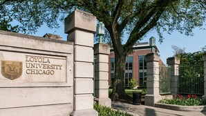 La entrada de la Universidad de Loyola Chicago con un cartel de piedra, un árbol grande y un edificio de ladrillo al fondo.