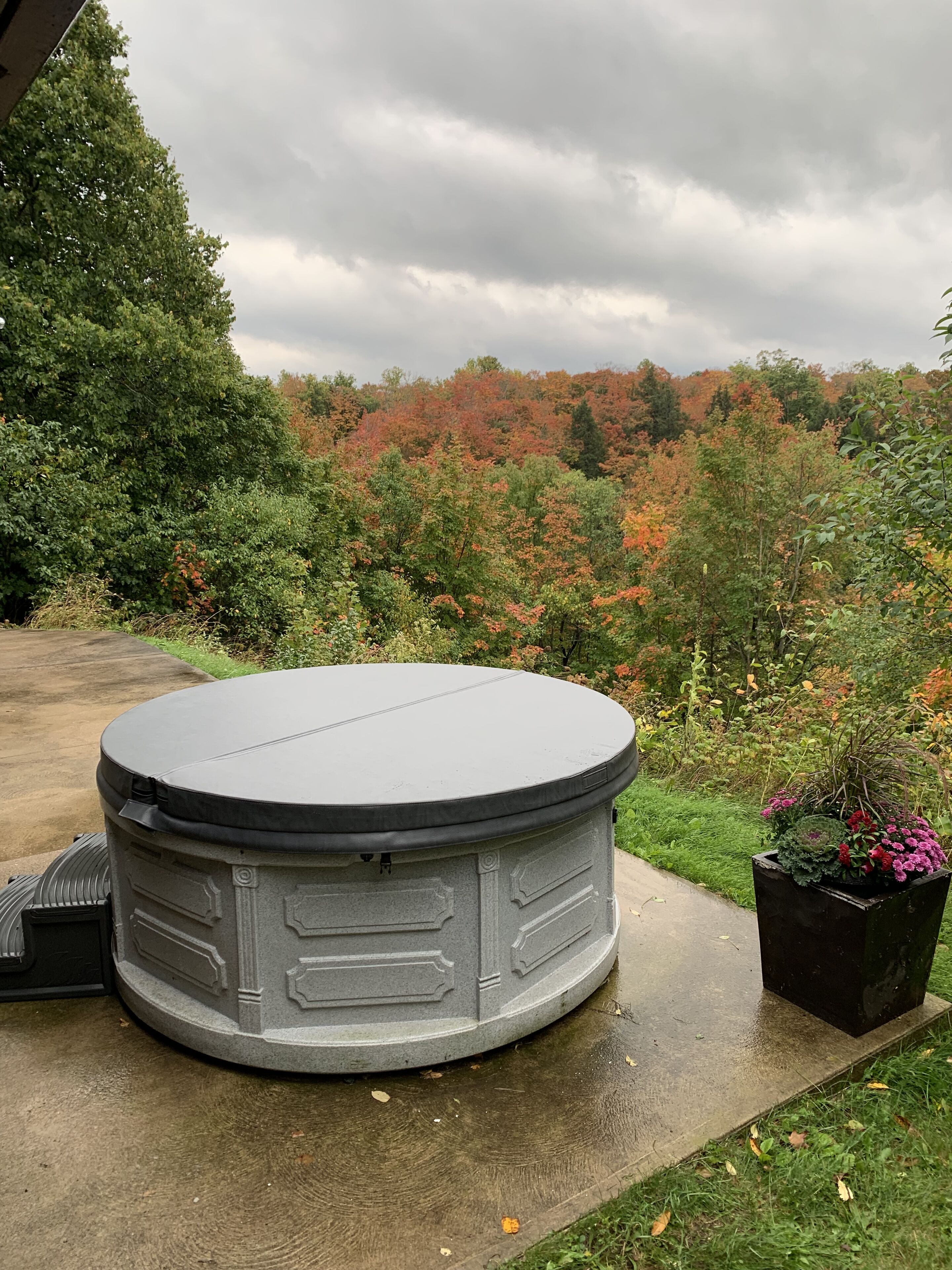 A hot tub with a stone exterior, surrounded by a garden with various plants and a view of a forested area.