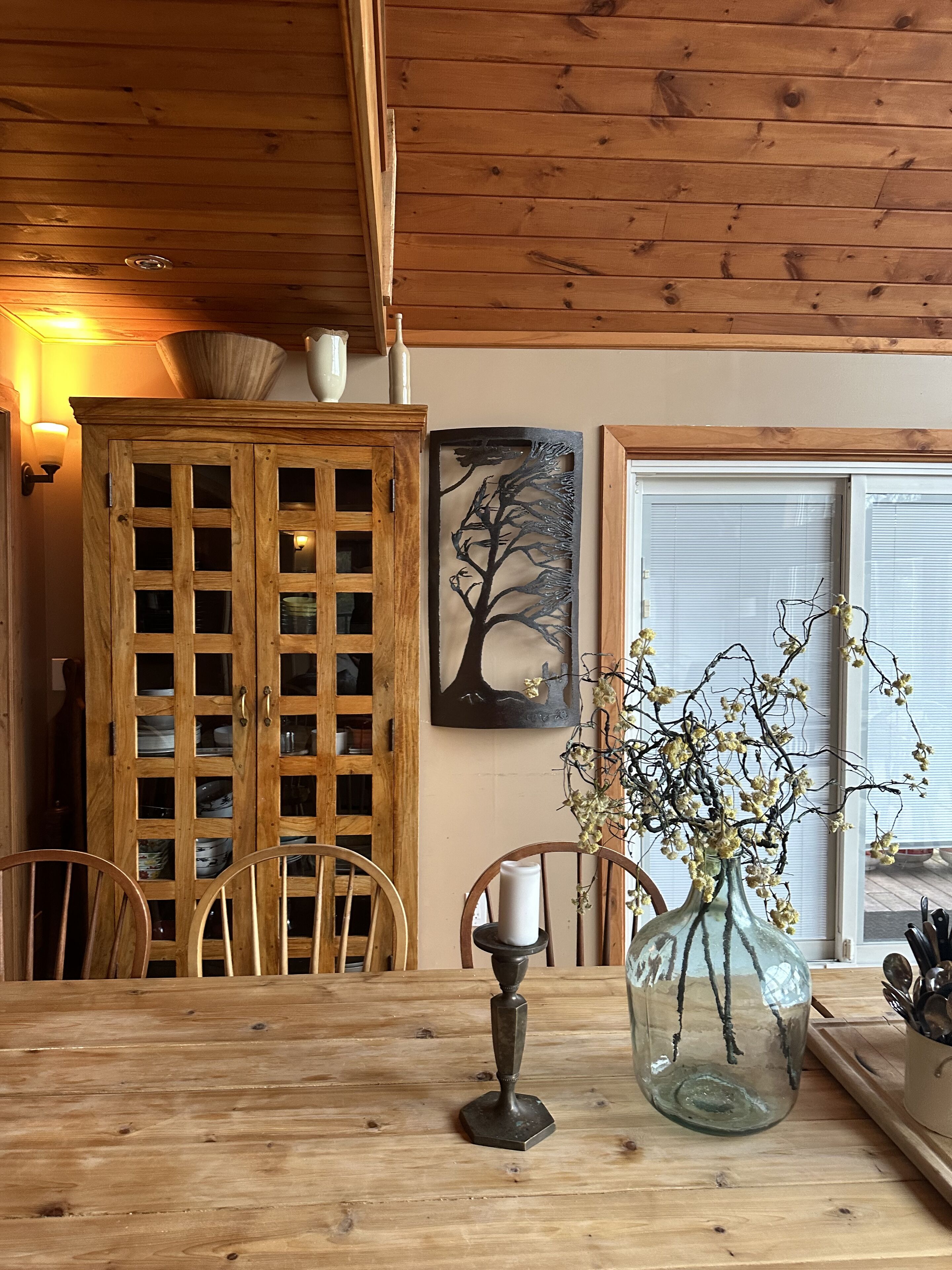 Une table à manger en bois avec des chaises, un meuble avec des portes en verre, un tableau accroché au mur et un vase décoré de branches.