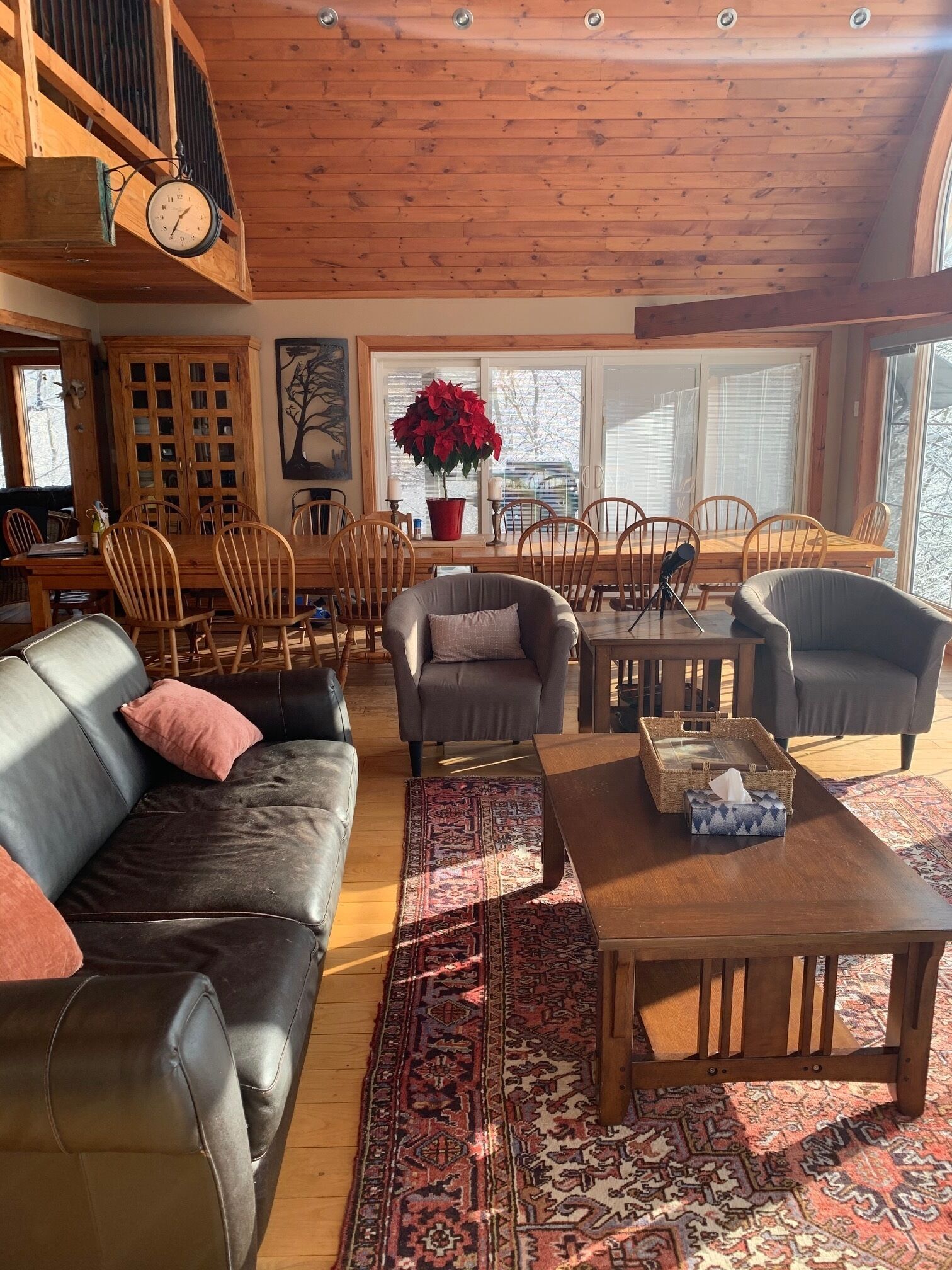 A cozy living room with a wooden ceiling, a dining area with wooden chairs, a leather sofa, and a large window with a view.