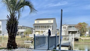 A pathway leading to a building with a balcony, surrounded by a palm tree and a small water body.