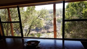 A dining table with a bowl in the center, large windows with a view of trees and a river. - Secluded Waterfront Cabin- Canoe, Fish, Swim & Stargaze near Pedernales Falls (Dripping Springs)