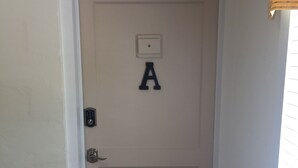 Eine weiße Tür mit einem Hinweisschild mit dem Buchstaben 'A' darüber, einem Klingel und einem Fenster mit Jalousien zur Rechten.