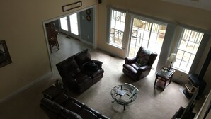 A spacious living room with a large brown leather sofa, a smaller brown leather chair, and a glass coffee table. There is a fireplace on the right and a view of the backyard through large windows. - Great waterfront relaxation! (Bumpass)