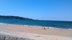 A beach with a clear sky, calm sea, and a distant hill with buildings.