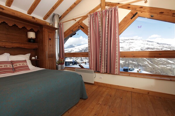 Une chambre avec un grand lit, des poutres en bois, une fenêtre avec des rideaux, un radiateur et une vue sur des montagnes enneigées.