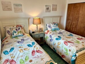 A bedroom with two beds, each with a beach-themed bedspread. There is a bedside table with a lamp and a small decorative item.