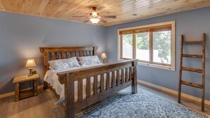 Ein Schlafzimmer mit einem hölzernen Bett, einem Nachttisch mit Lampe, einem Fenster mit Ausblick und einer Holzleiter.