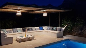 Ett takförsett utomhusområde med sittplatser och en pool, inrett med en vit soffa med mönstrade kuddar, ett soffbord och hängande lampor.