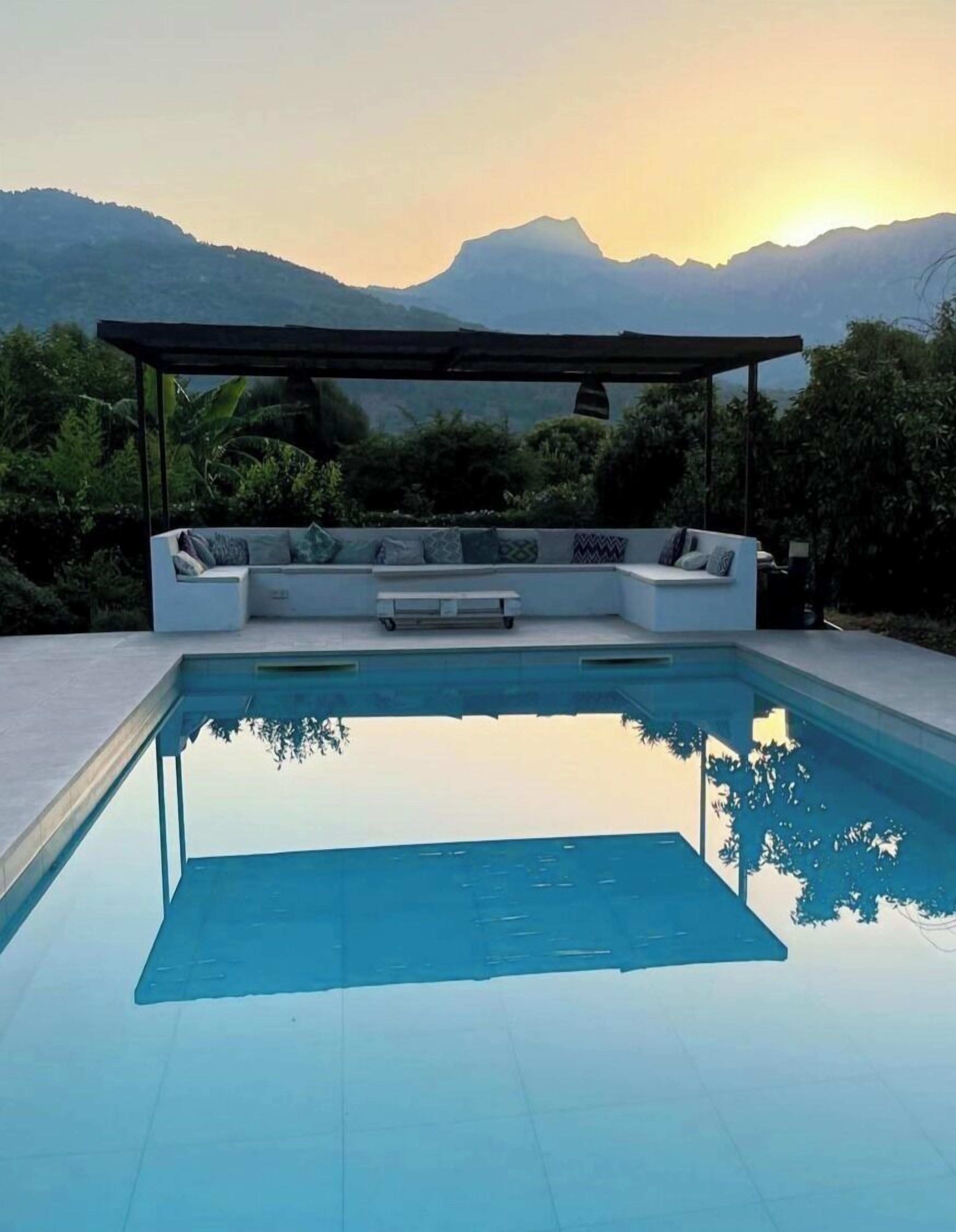 A swimming pool with a seating area and a pergola, surrounded by greenery and mountains.
