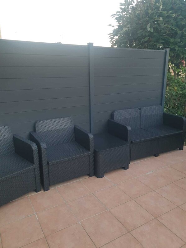 Una fila de asientos de mimbre oscuro para exteriores con un suelo de patio cubierto de baldosas.