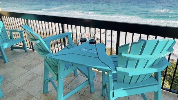 Un espace extérieur aménagé en bleu turquoise, avec une table et des chaises, donnant sur une plage où l’on peut admirer les vagues et un ciel dégagé.