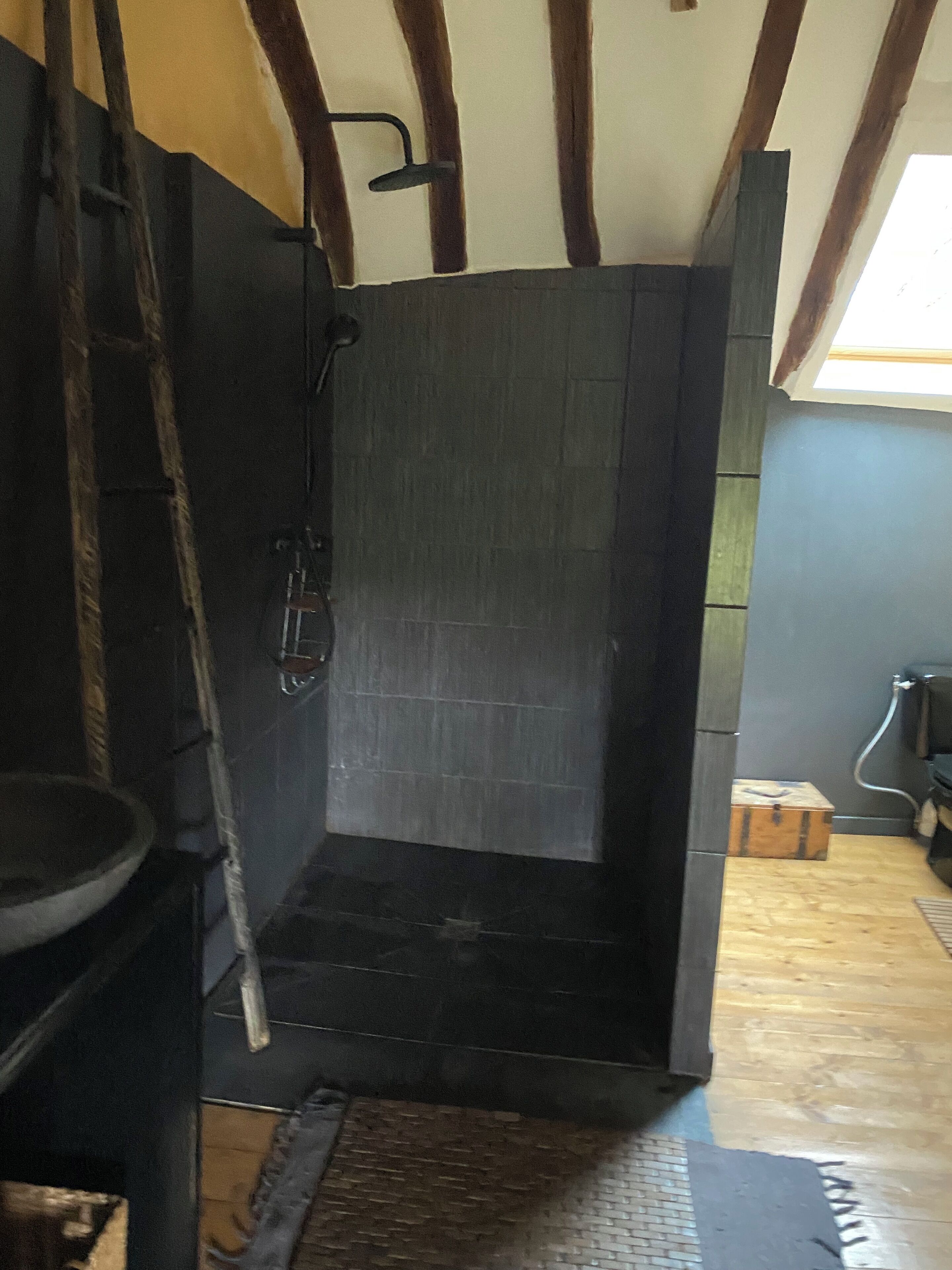A bathroom with a dark tiled shower, a wooden ladder, a small wooden chest, and a window with natural light.