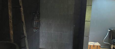 A bathroom with a dark tiled shower, a wooden ladder, a small wooden chest, and a window with natural light.