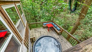 Una terraza de madera con jacuzzi, zona de estar y amplios ventanales con vistas a un área boscosa.