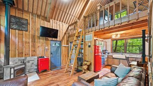 Interior rústico de una cabaña con paredes de madera, una chimenea de piedra, un refrigerador rojo, una escalera de madera y un dormitorio en entrepiso con ventana que da a la vegetación.