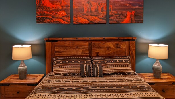 Un dormitorio con cabecera de madera, ropa de cama estampada, dos lámparas de noche y una pared decorada con tres cuadros de paisajes.