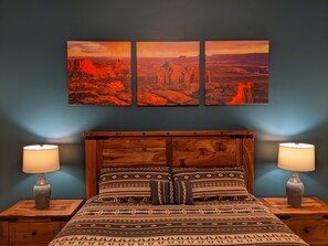 Ein Schlafzimmer mit einem Holzkopfteil, gemusterten Betttextilien, zwei Nachttischlampen und einer Wand mit drei Landschaftsbildern.